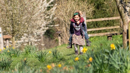 A girl carrying a little boy on her back while smiling and walking through daffodils and tall grass. There are some blossoming orchard trees around them.